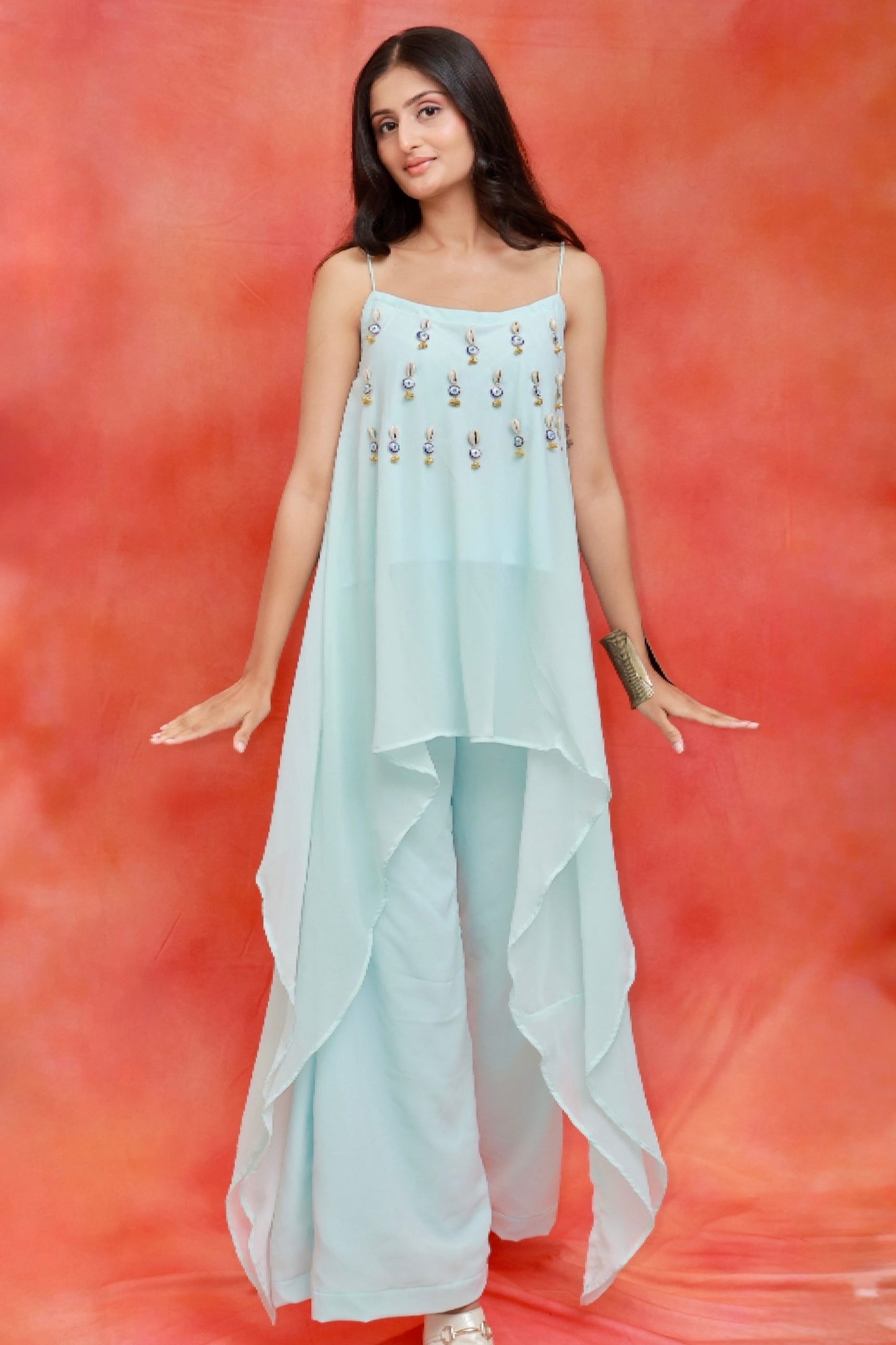 Model wearing the Nazar Ice Blue Shell Co-ord Set in premium georgette featuring shell and evil-eye embellishments, an asymmetrical layered camisole top, and matching wide-leg pants.