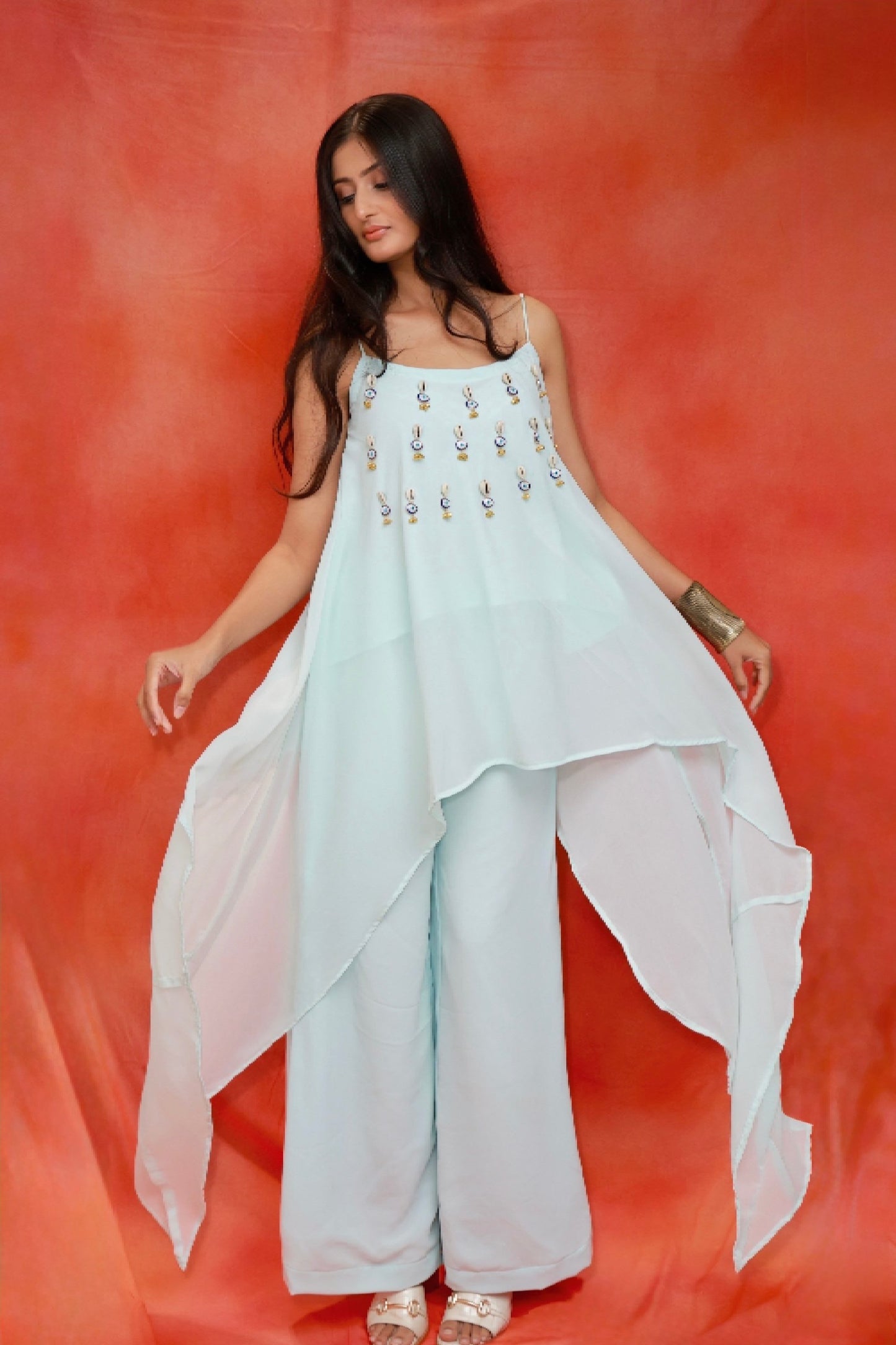 Model wearing the Nazar Ice Blue Shell Co-ord Set in premium georgette featuring shell and evil-eye embellishments, an asymmetrical layered camisole top, and matching wide-leg pants.